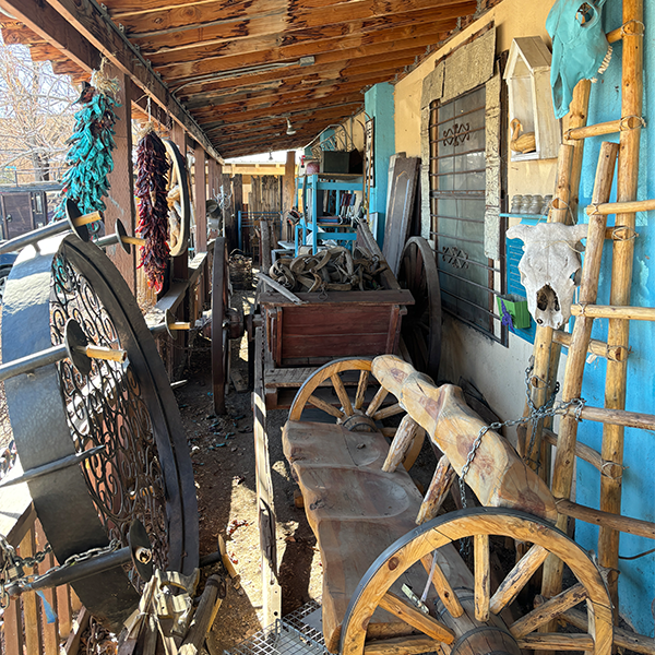 A crowded porch features many interesting items for sale, including a bench made out of wagon wheels and driftwood, native american-style wood ladders, cow skulls, a chandelier and a blue chili ristra.