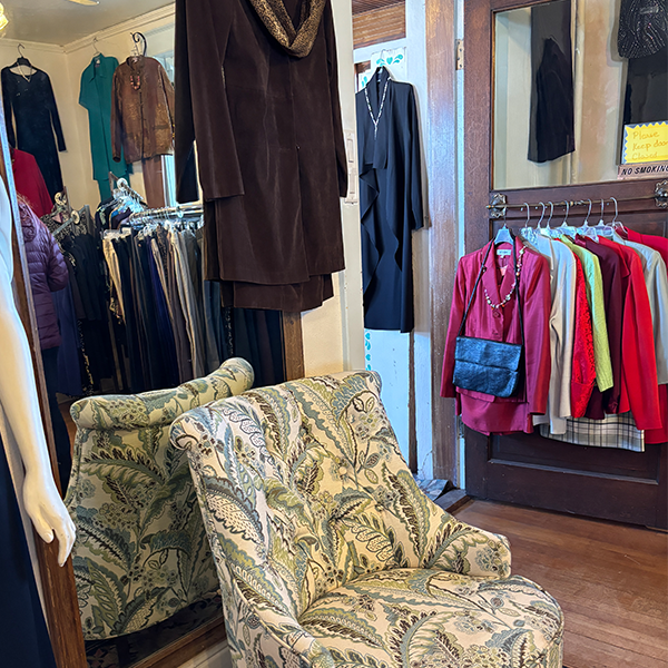 A nice armchair in front of a large mirror is surrounded by suit jackets, purses and pearls. It looks ready for someone to step out of the dressing room.